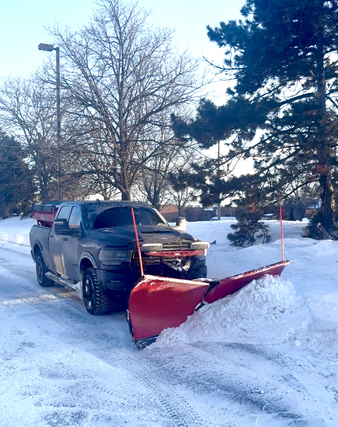 Black pickup truck with a red snow plow attached to the front, clearing snow on a snowy road during daytime