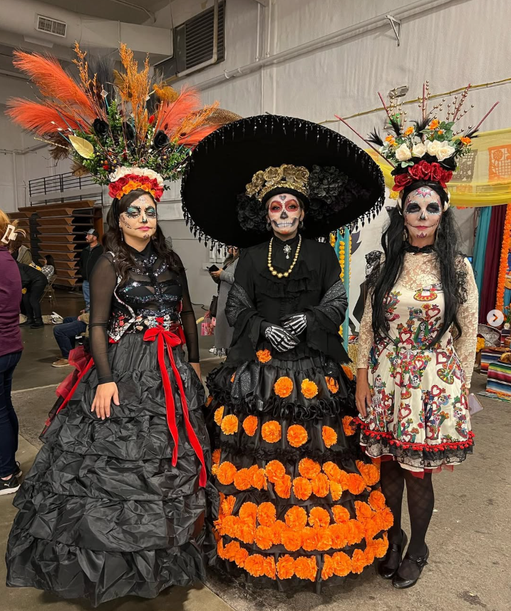Three women dressed in elaborate Día de los Muertos costumes, with sugar skull face paint and vibrant headpieces, standing indoors at a festive event.