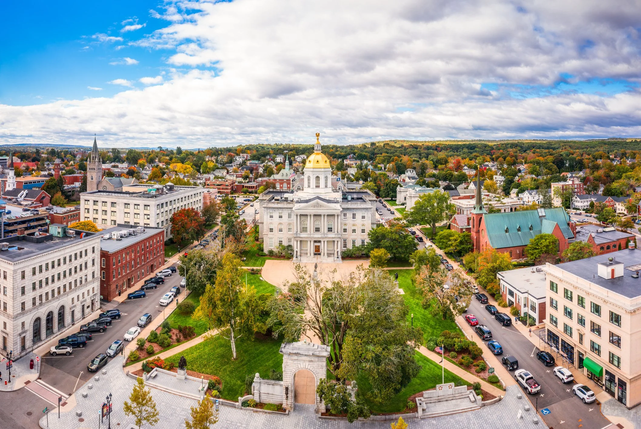 Aerial view of a town with historic buildings and a large white church with a gold dome in the center, surrounded by colorful trees and streets under a partly cloudy sky.