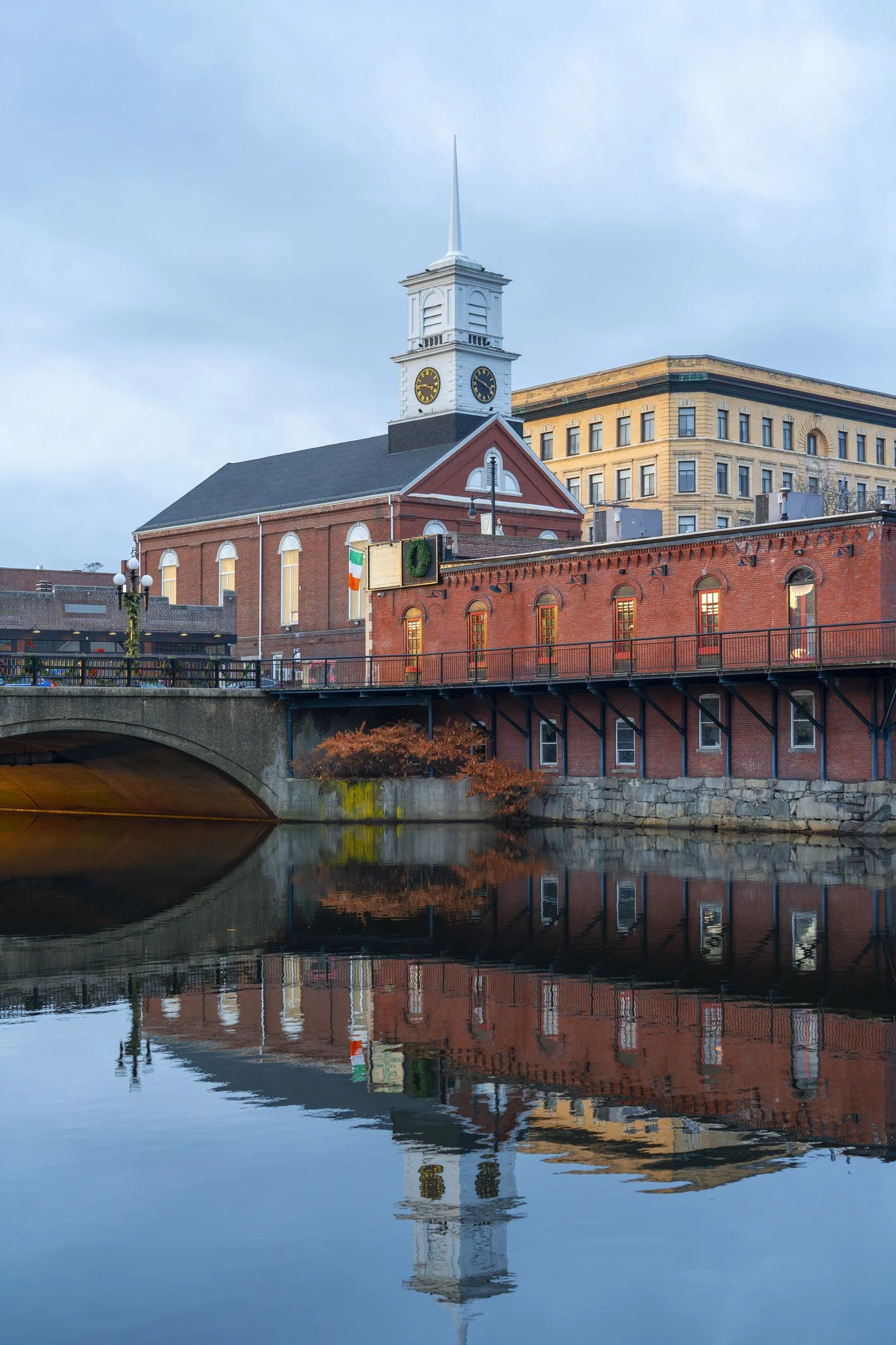 A historic brick building with a clock tower reflected in a calm river during dusk, with an overcast sky.