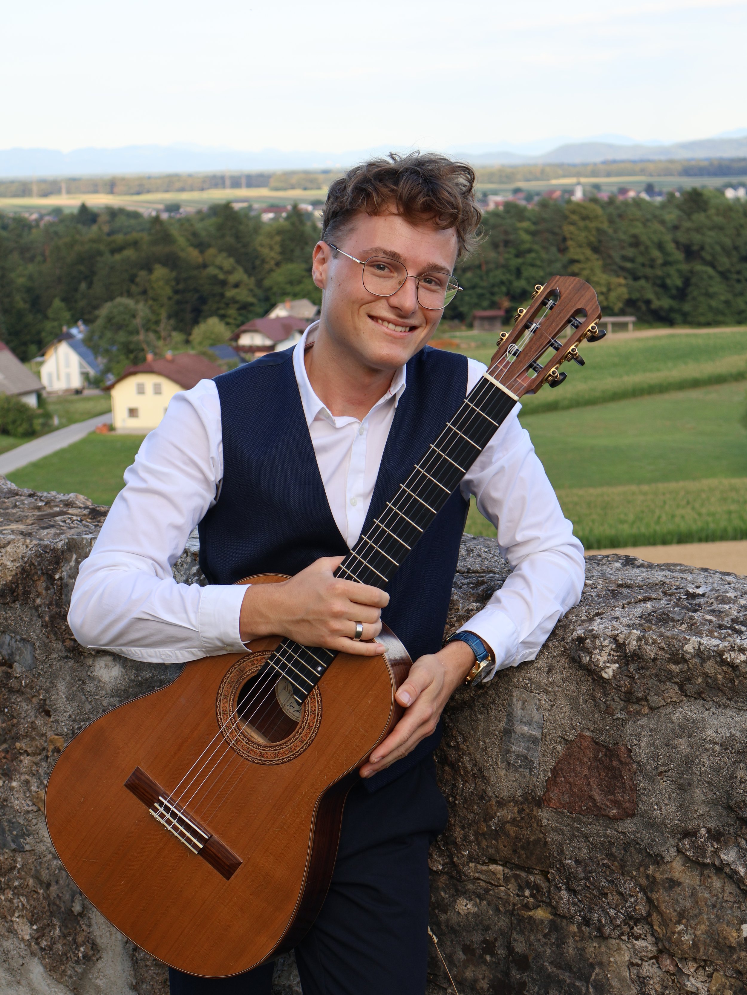A young man holding a guitar outdoors smiling at the camera with a rural landscape background.