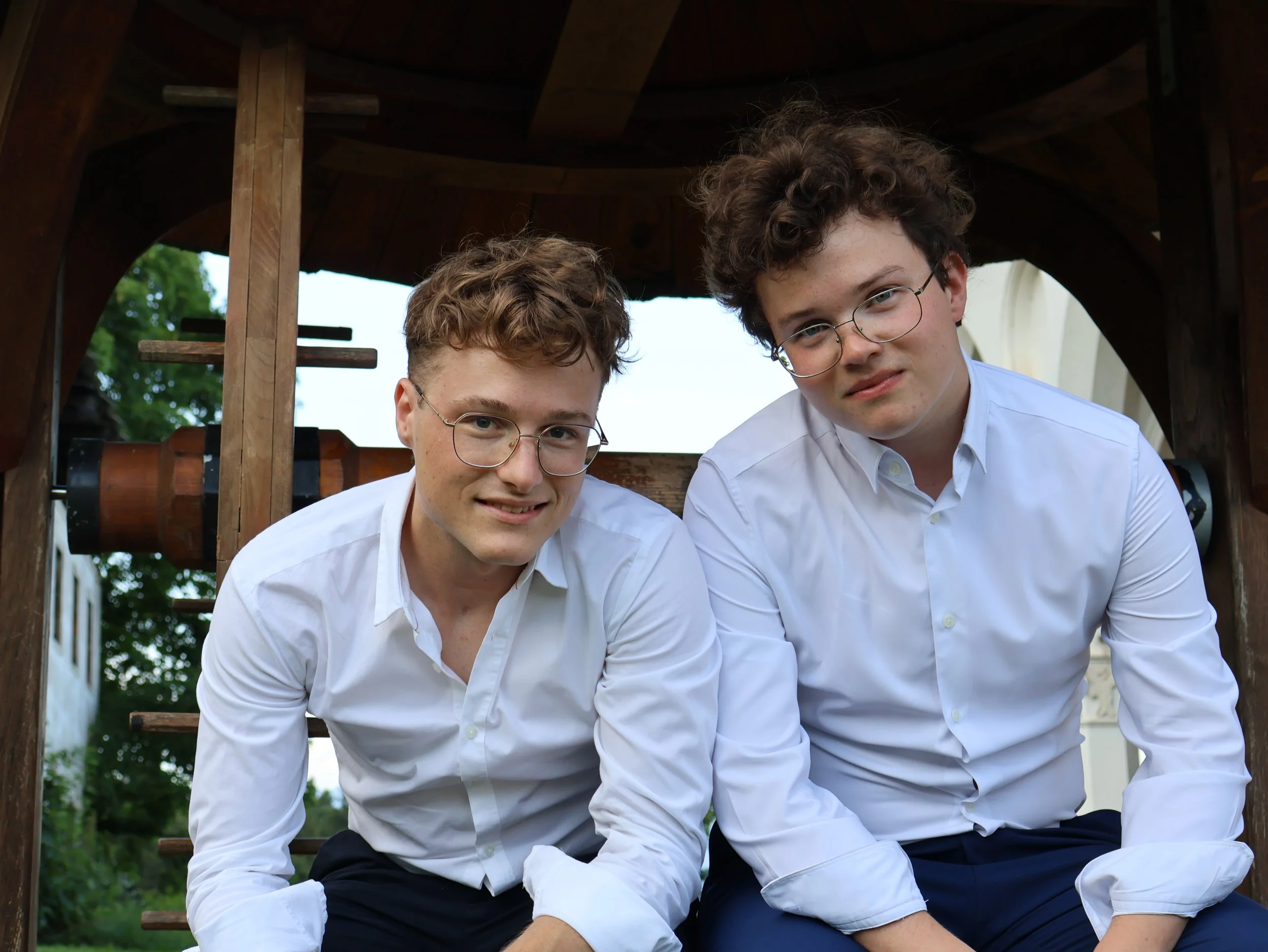 Two young men with glasses wearing white shirts and dark pants, sitting and leaning forward outdoors near a wooden structure with a large tube behind them.