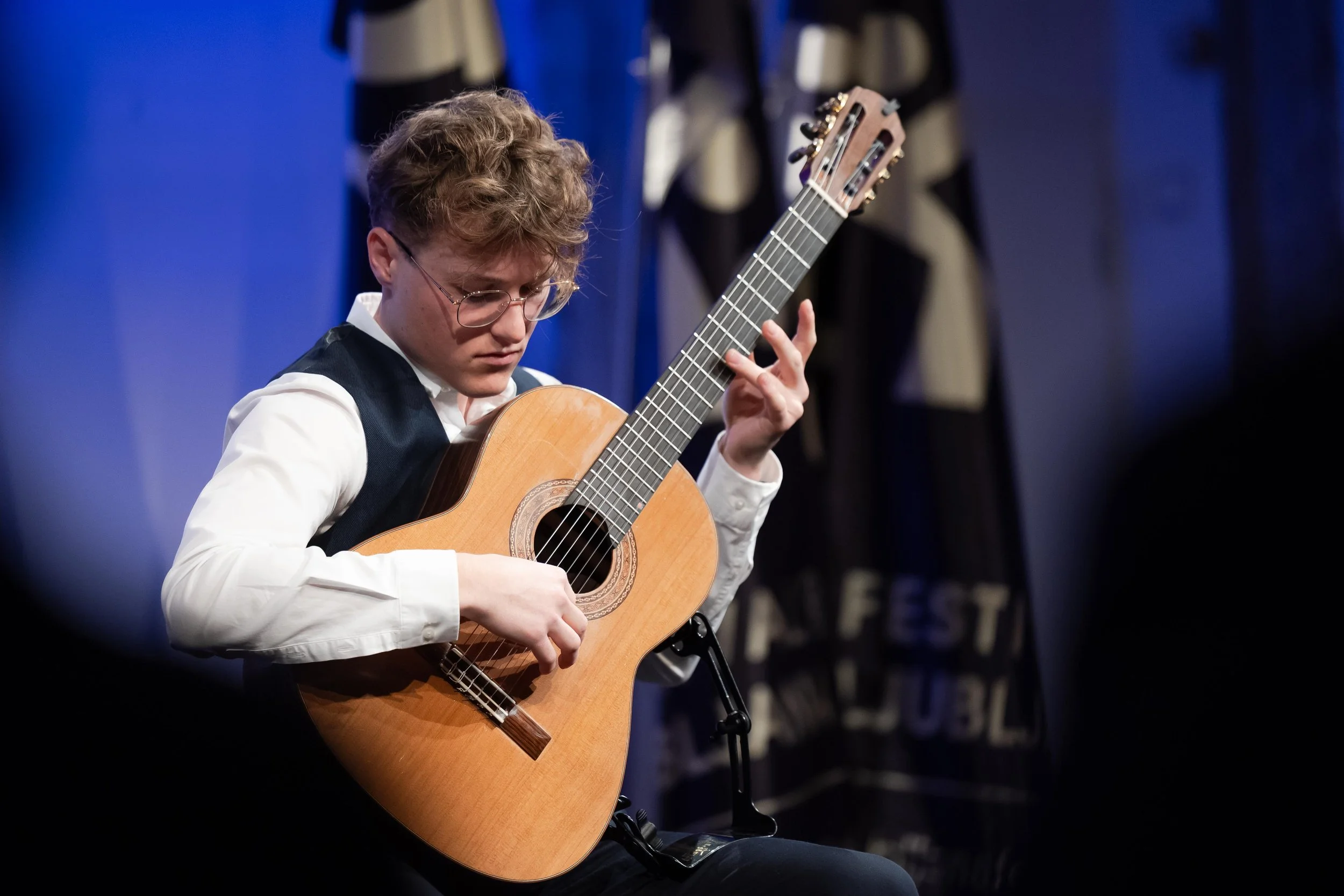 A young man with curly hair and glasses playing an acoustic guitar on stage during a performance.