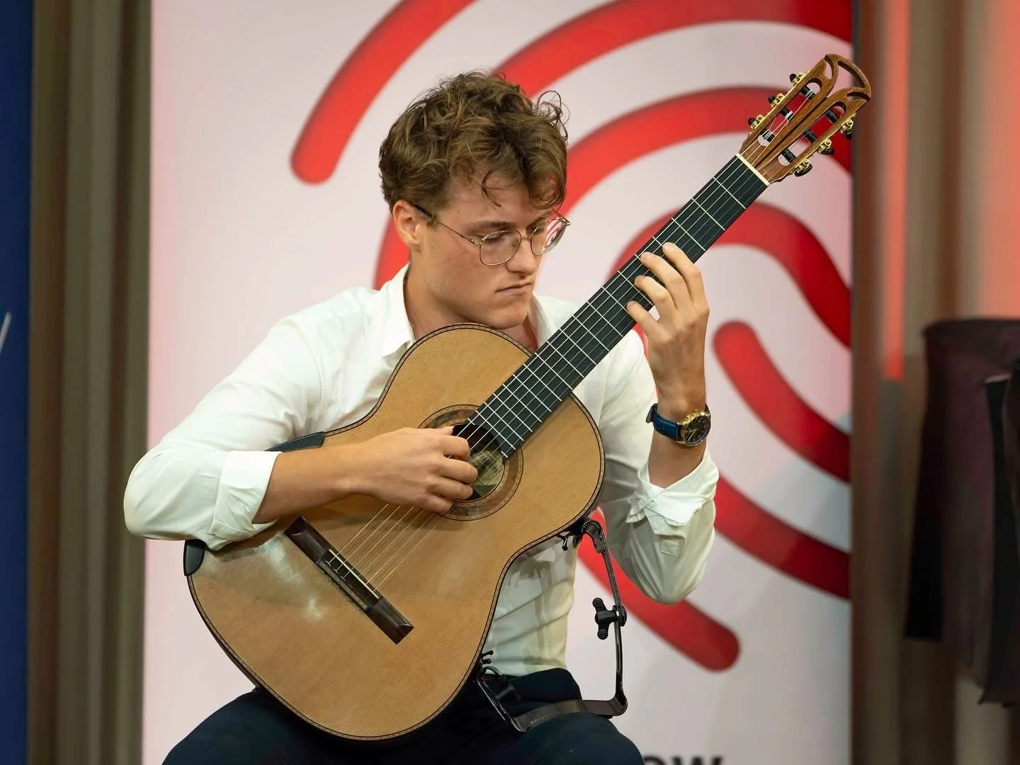 Young man playing classical guitar in front of a red and white abstract backdrop.