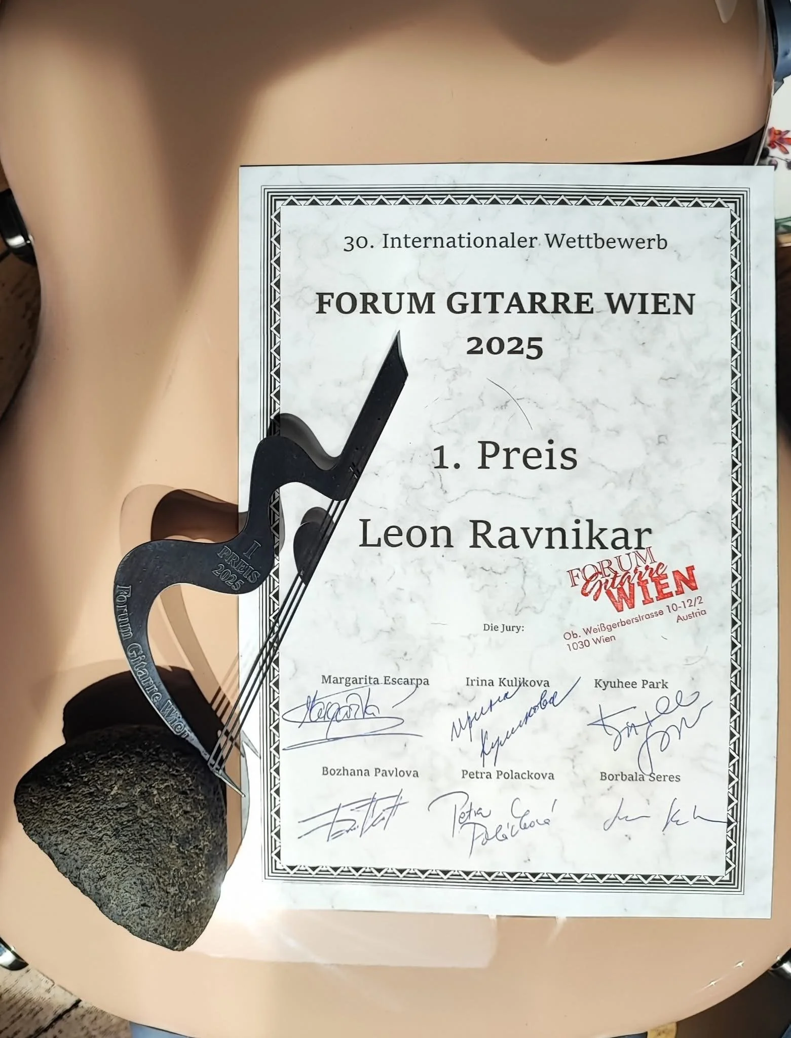 A guitar-themed trophy and a certificate from the 30th International Guitar Competition in Vienna, 2025, awarded to Leon Ravnikar for first place. The trophy is shaped like a guitar and is placed on a rock-like base, and the certificate features sign
