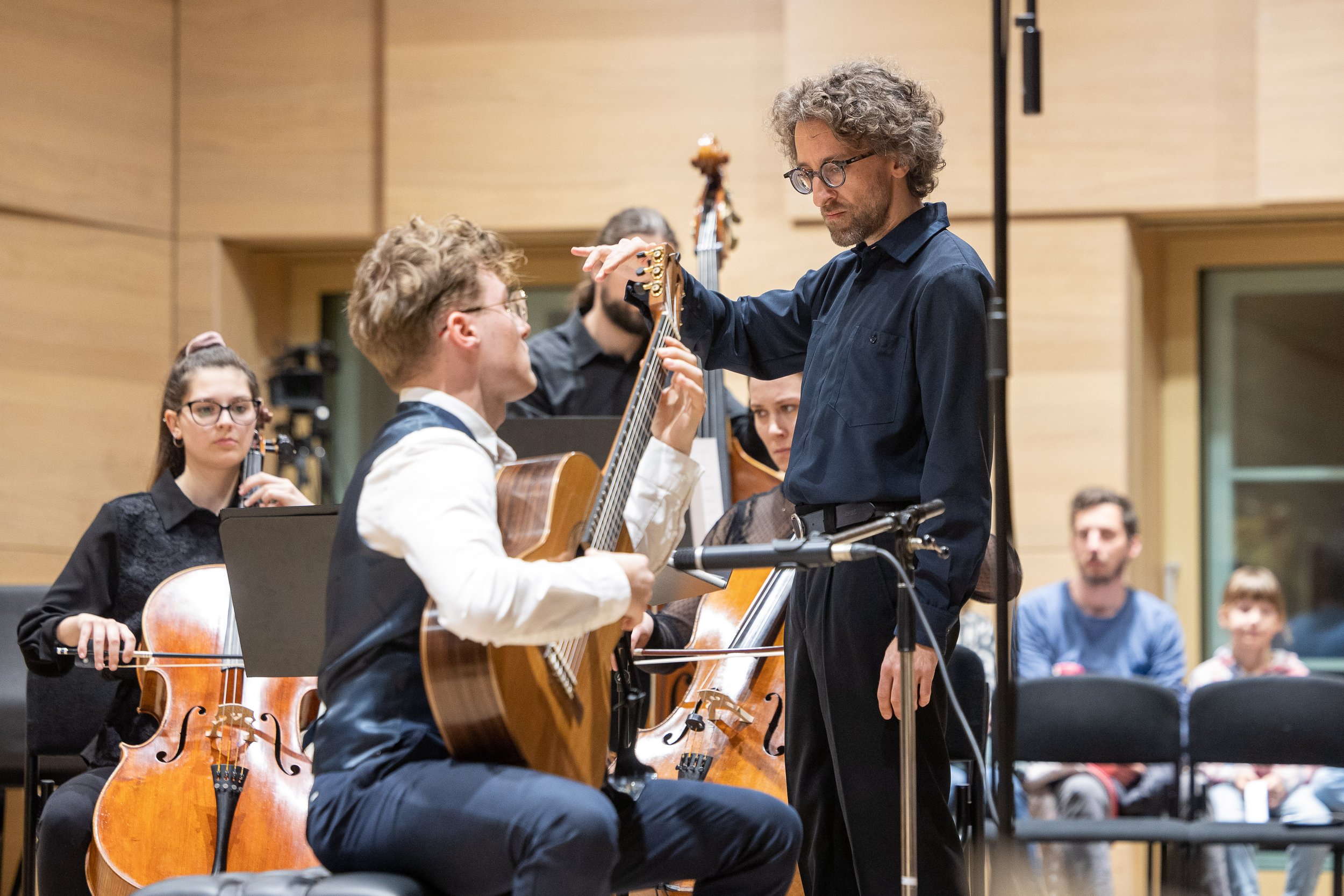 A group of musicians in a recording studio or concert hall, with a man conducting or giving instructions to a young guitarist. The young guitarist is seated, playing an acoustic guitar, while other musicians hold string instruments like violins and c