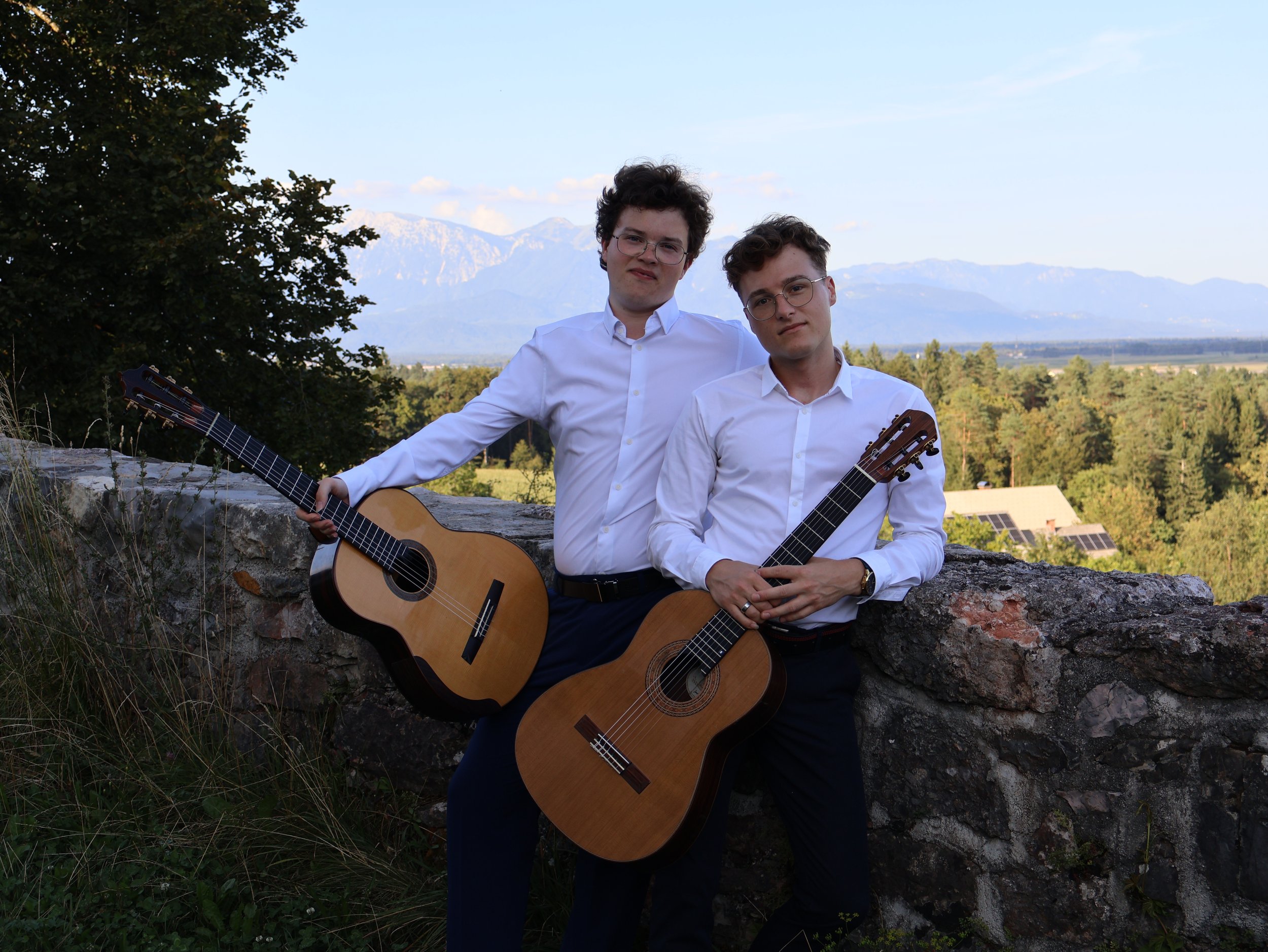 Two young men in white shirts holding guitars outdoors with a scenic mountain and forest background.