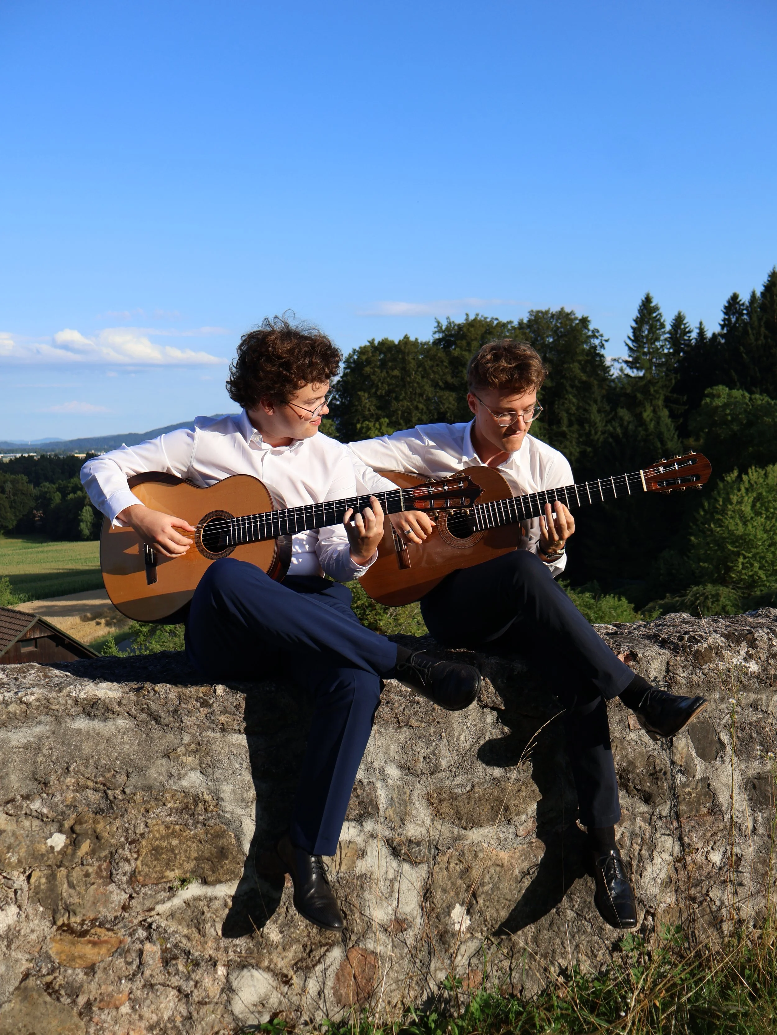 Two men sitting on a stone wall playing acoustic guitars outdoors, with trees and a blue sky in the background.