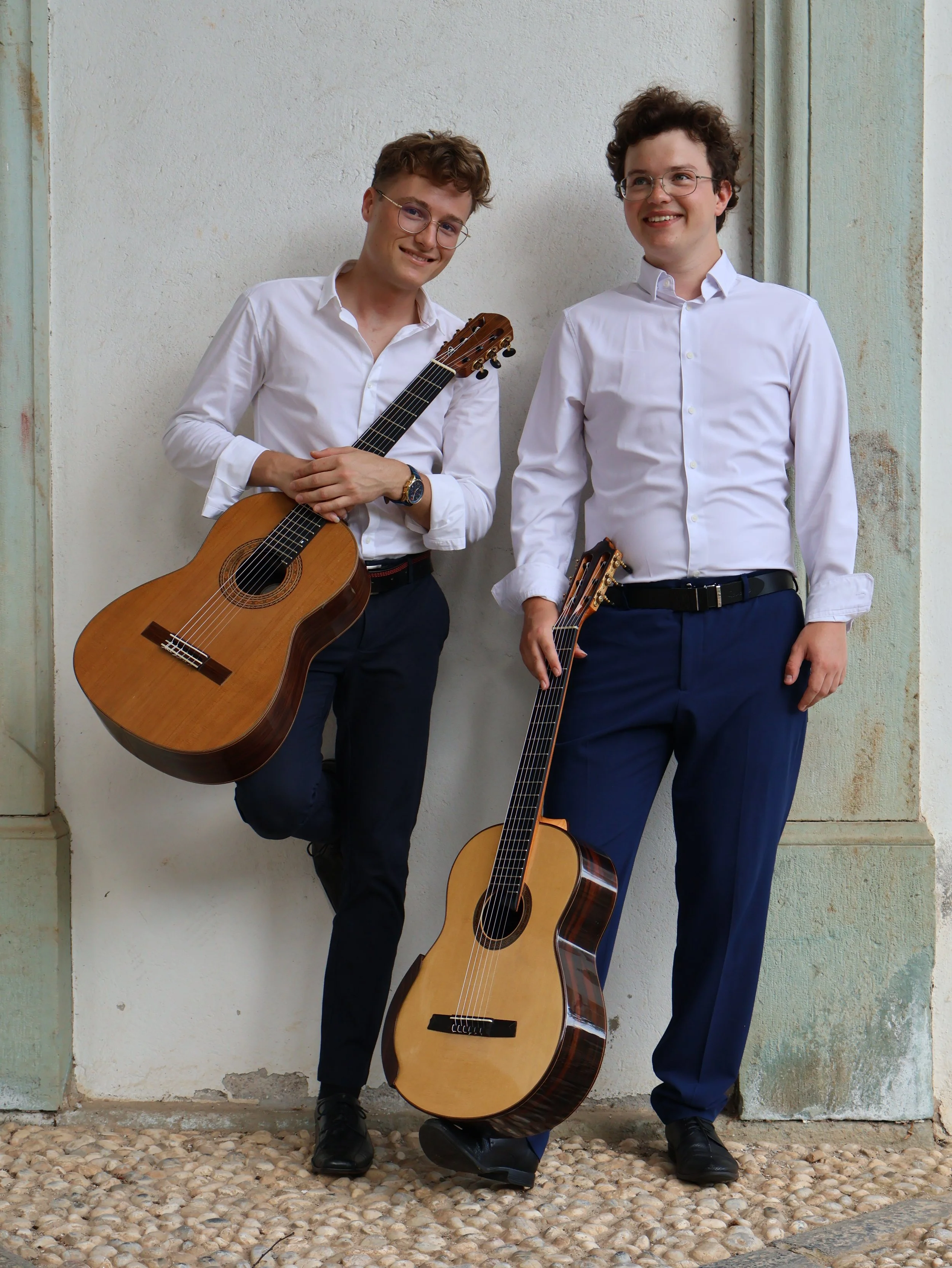 Two young men in white shirts and dark pants, holding guitars, standing against a textured wall and smiling.