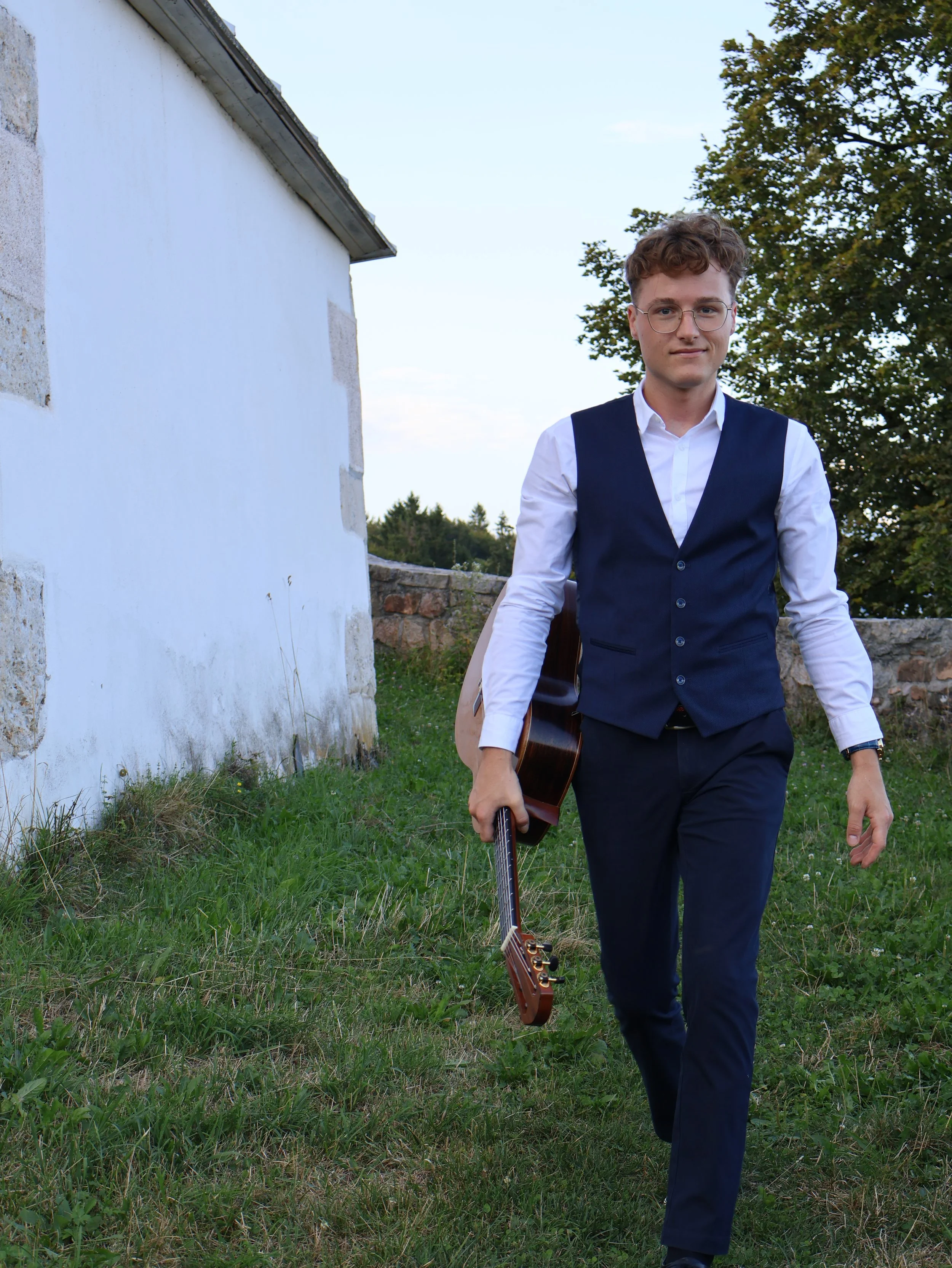 A young man in formal attire walking outdoors on grass, holding a guitar in his right hand, with a white building and trees in the background.