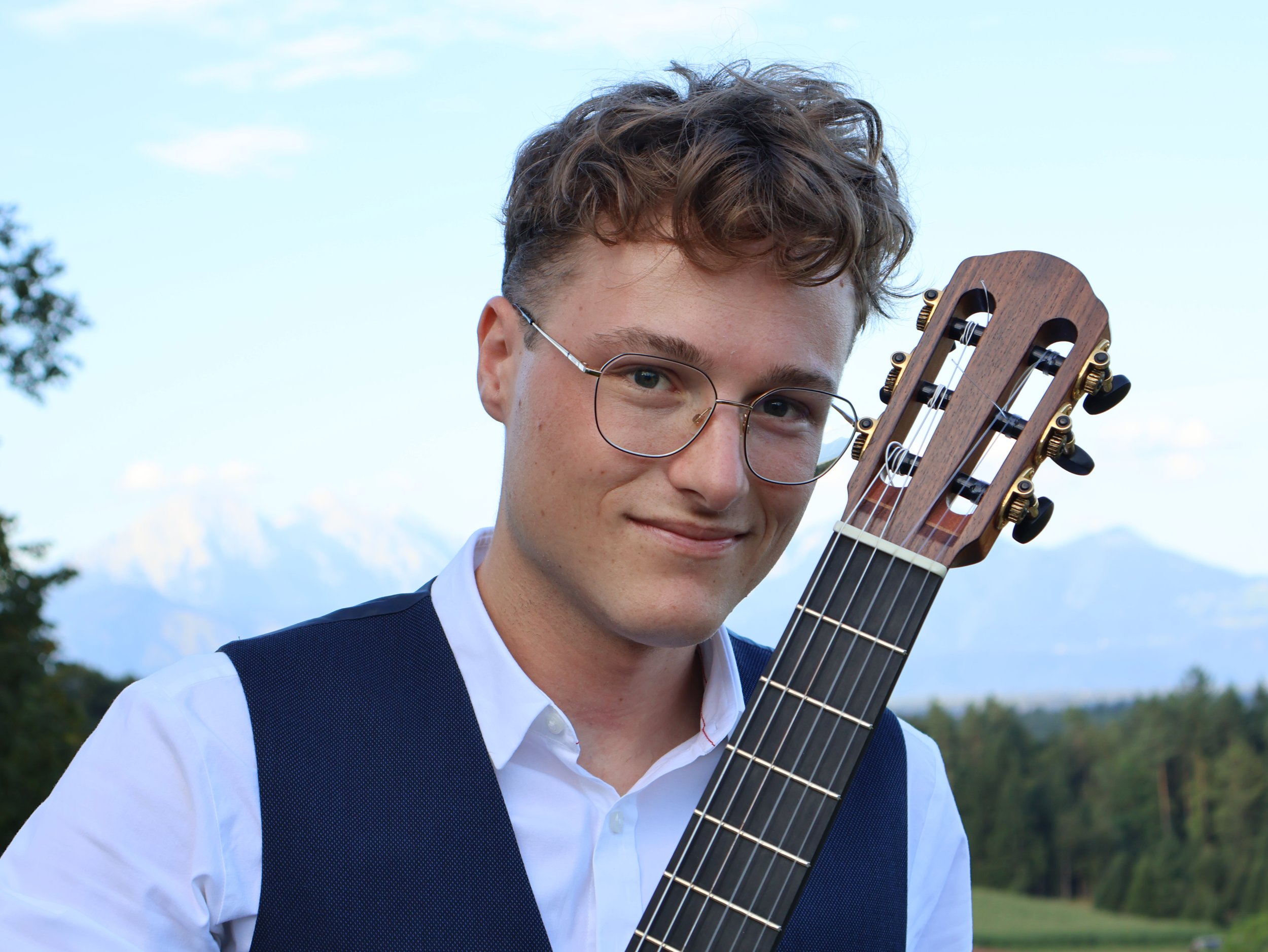 Young man with curly hair and glasses smiling outdoors while holding a wooden acoustic guitar.