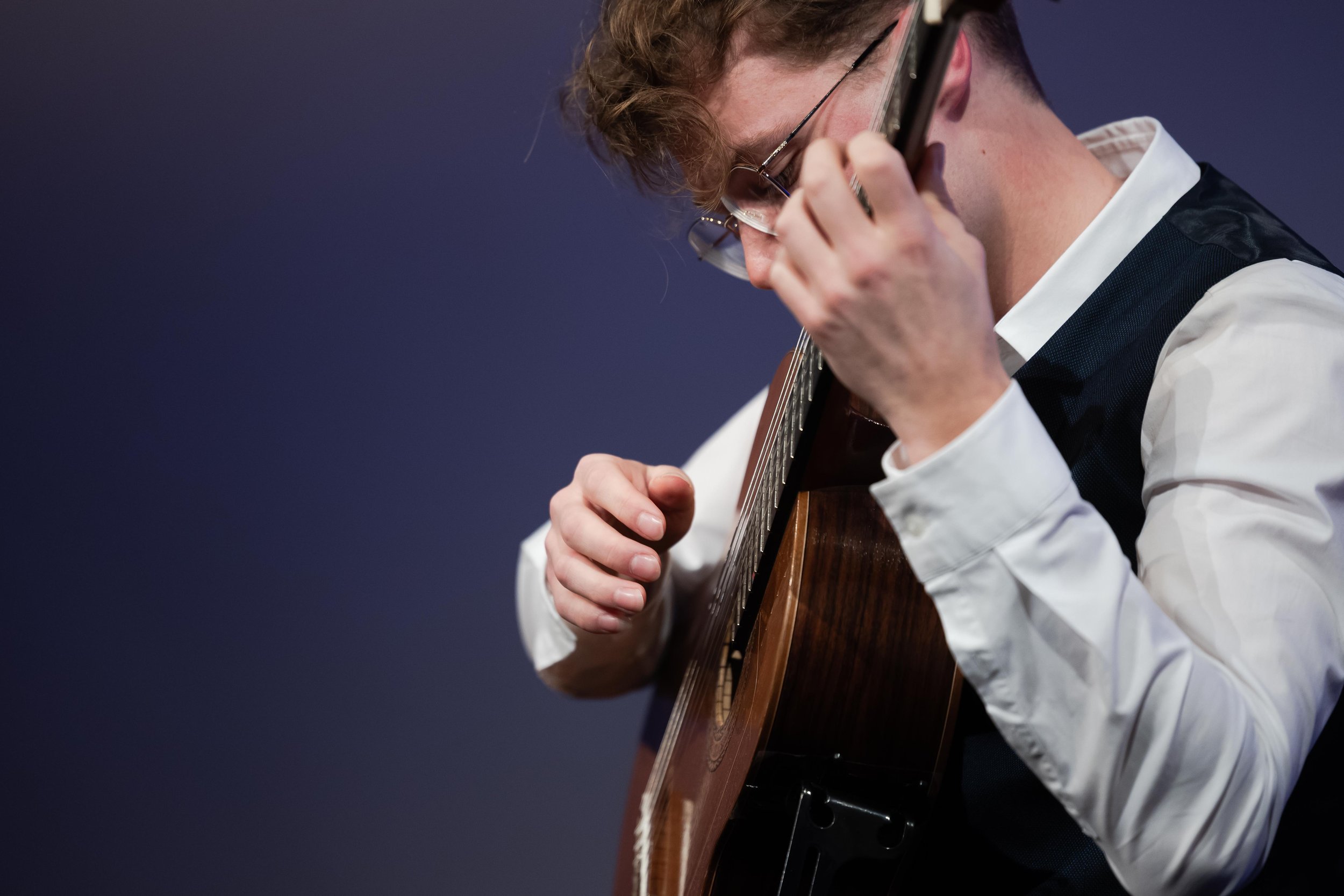 A young man with curly hair, glasses, dressed in a white shirt and black vest, playing an acoustic guitar.