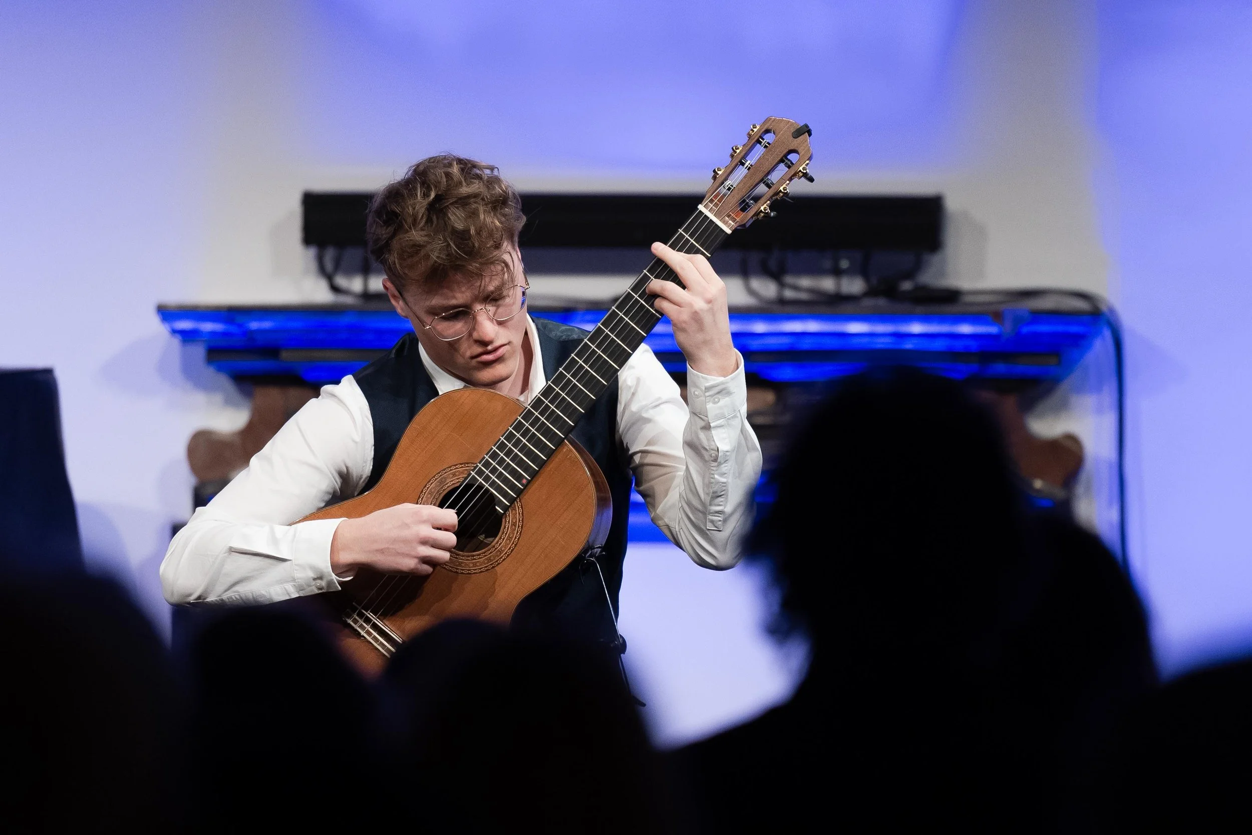 Young man with curly hair and glasses playing classical guitar during a performance, with a blue-lit stage and audience in the foreground.