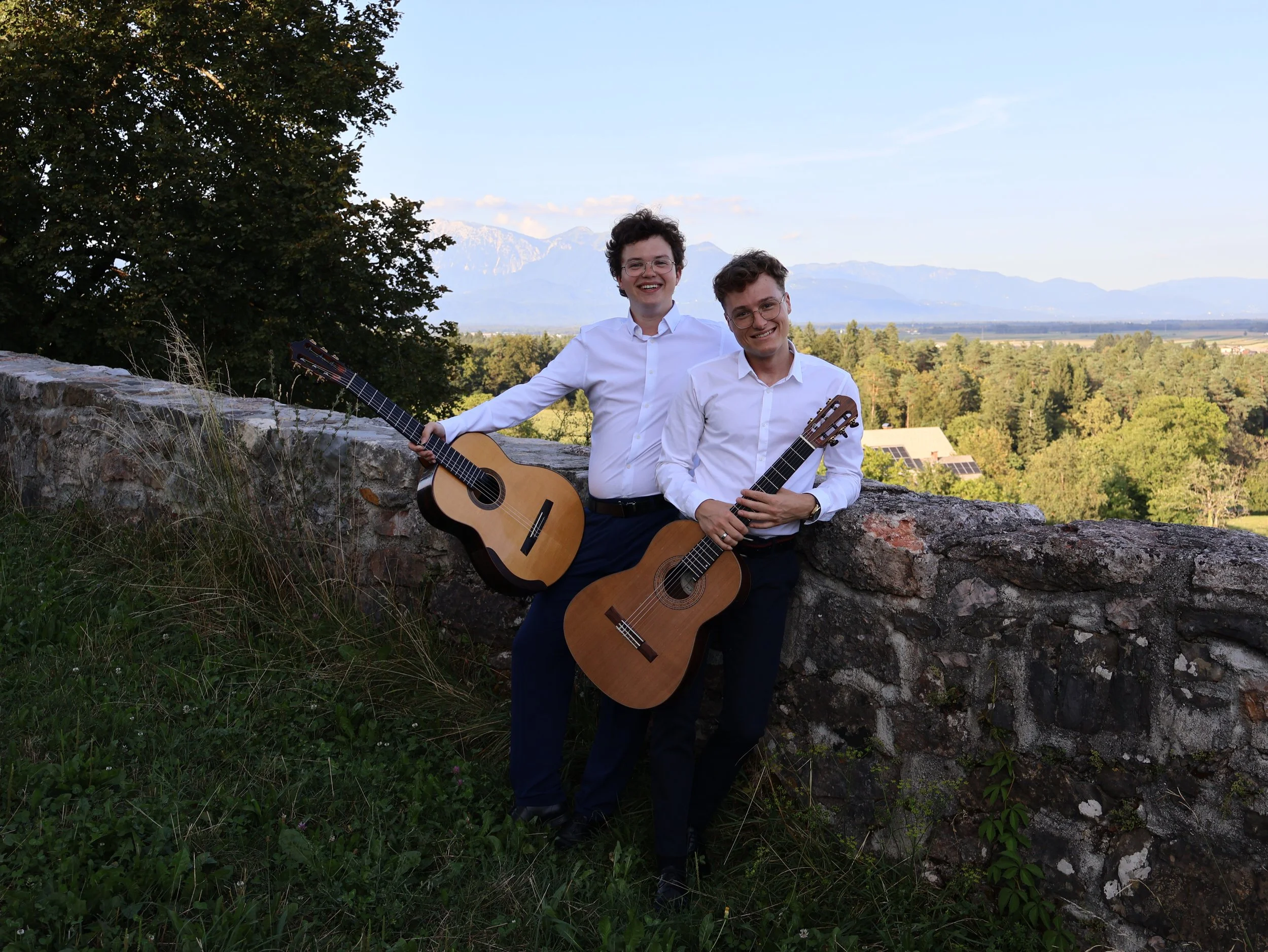 Two young men dressed in white shirts and dark pants, holding acoustic guitars, standing outdoors by a stone wall with a scenic view of trees, houses, and mountains in the background.