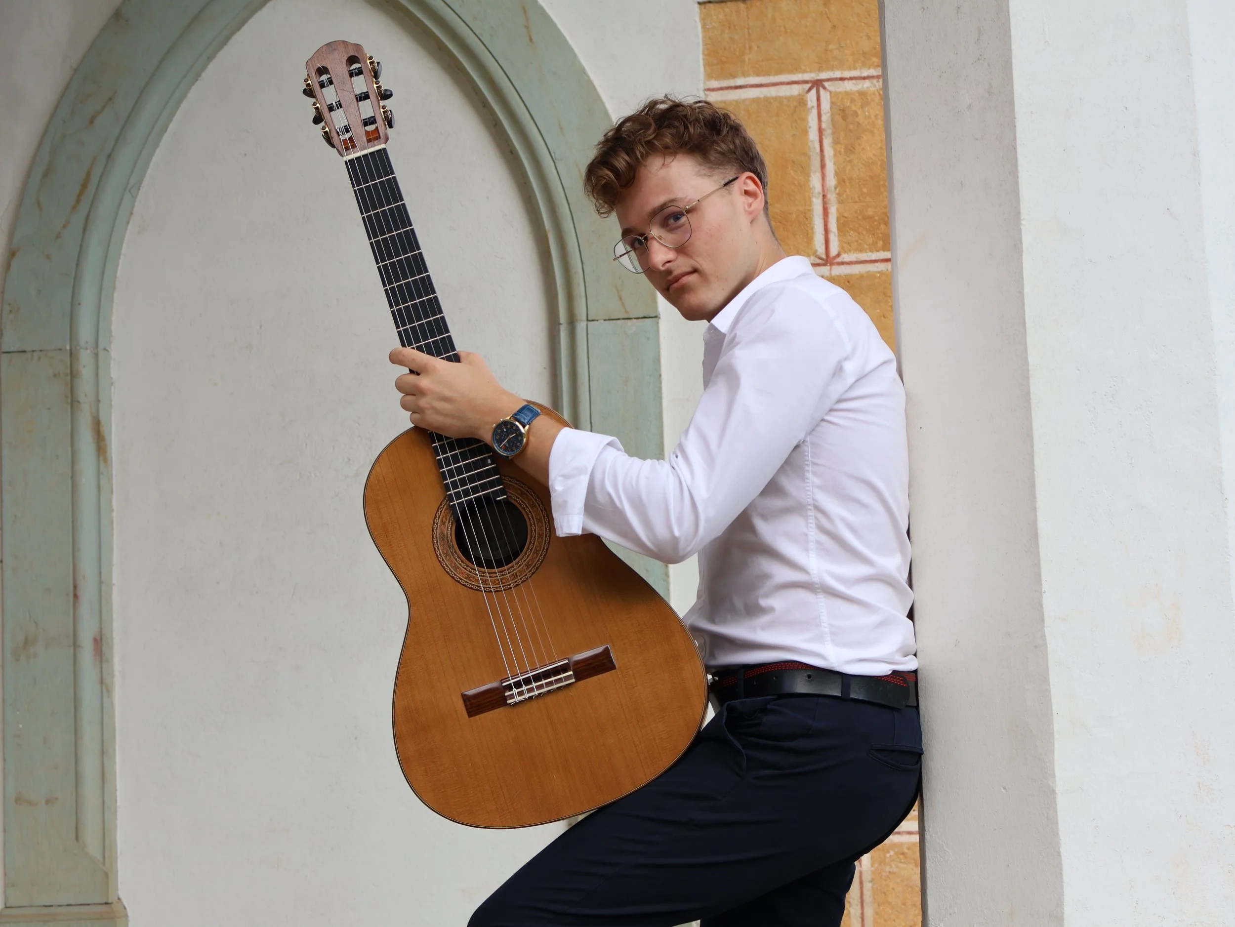 A young man with curly hair and glasses, wearing a white shirt, sitting and holding an acoustic guitar.
