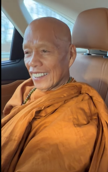 A smiling elderly Buddhist monk wearing traditional orange robes sitting in a car seat.
