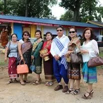 Five women standing outdoors in front of a colorful building, holding bags, with one man among them. They are dressed in casual and traditional clothing.