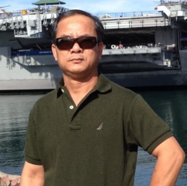 Man wearing sunglasses and a dark green polo shirt posing outdoors near water with a large ship in the background.