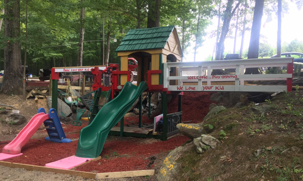 Children's playground with slides, swings, and a small playhouse, surrounded by trees and rocks, with signs that say 'Take care your kid,' 'Welcome Playground,' and 'We Love your Kid'.