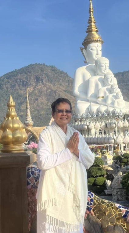 A woman in white traditional clothing standing with folded hands in front of a large white and gold multi-headed Buddha statue outdoors with a mountain in the background.