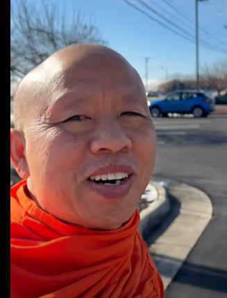 A smiling person with a shaved head wearing an orange jacket standing outside on a sunny day near a parking lot with cars and power lines in the background.