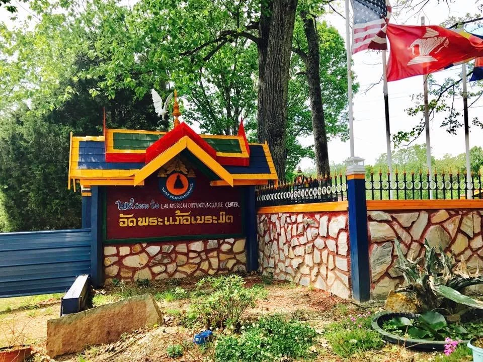 Entrance sign for the Lao American Community & Culture Center, surrounded by trees, with flags and a stone wall.