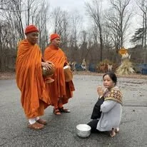 A woman kneels on the ground with a bowl in front of her, while two Buddhist monks in orange robes and red hats stand nearby holding alms bowls, outdoors with bare trees in the background.