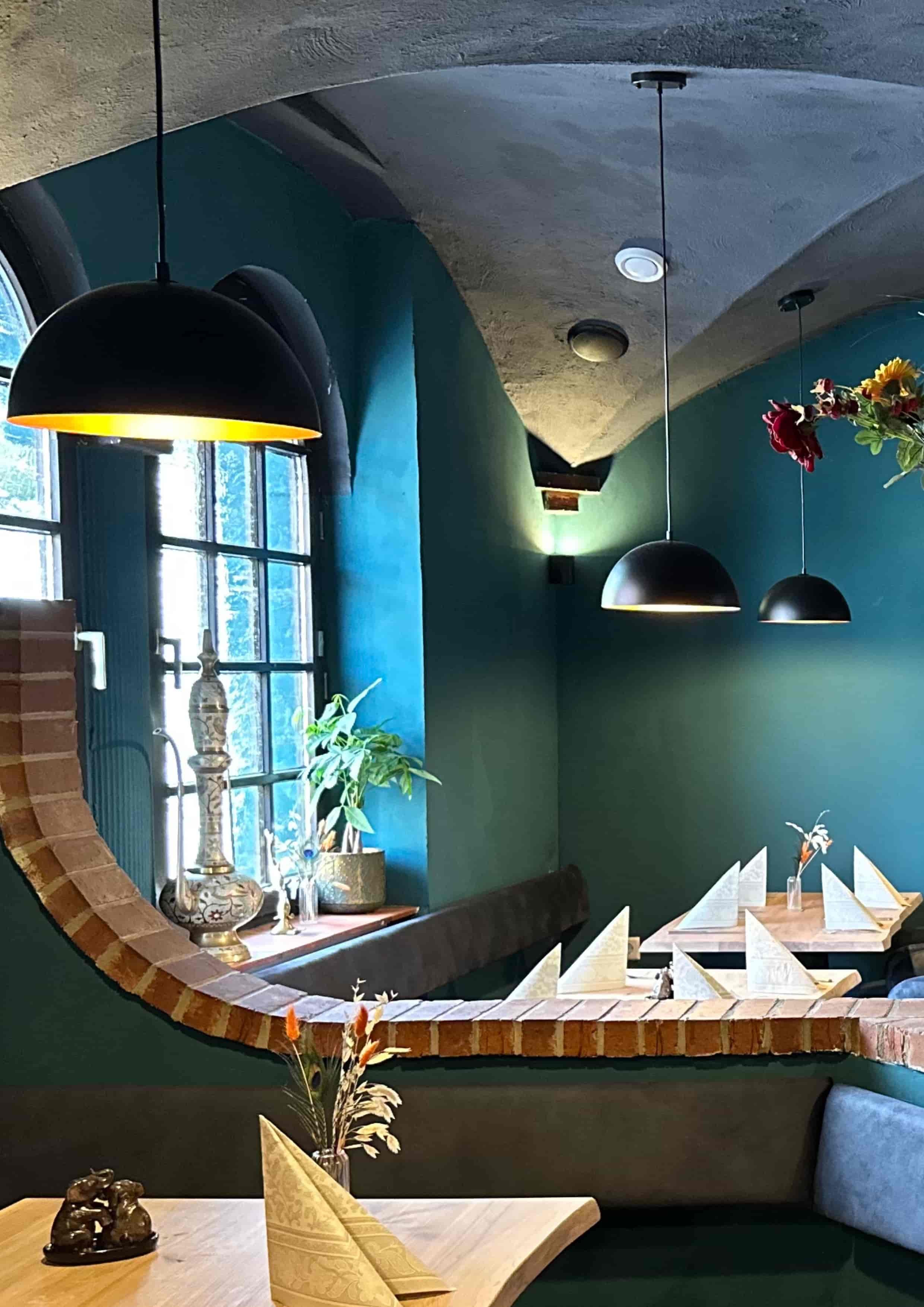 Ein Blick in ein elegantes Restaurant mit dunklen Wänden, künstlicher Beleuchtung und gedeckten Tischen mit weißem Geschirr. Im Vordergrund ein dekorativer Tisch mit einer Pflanze und einem Kerzenständer, im Hintergrund Kamine und Fenster.