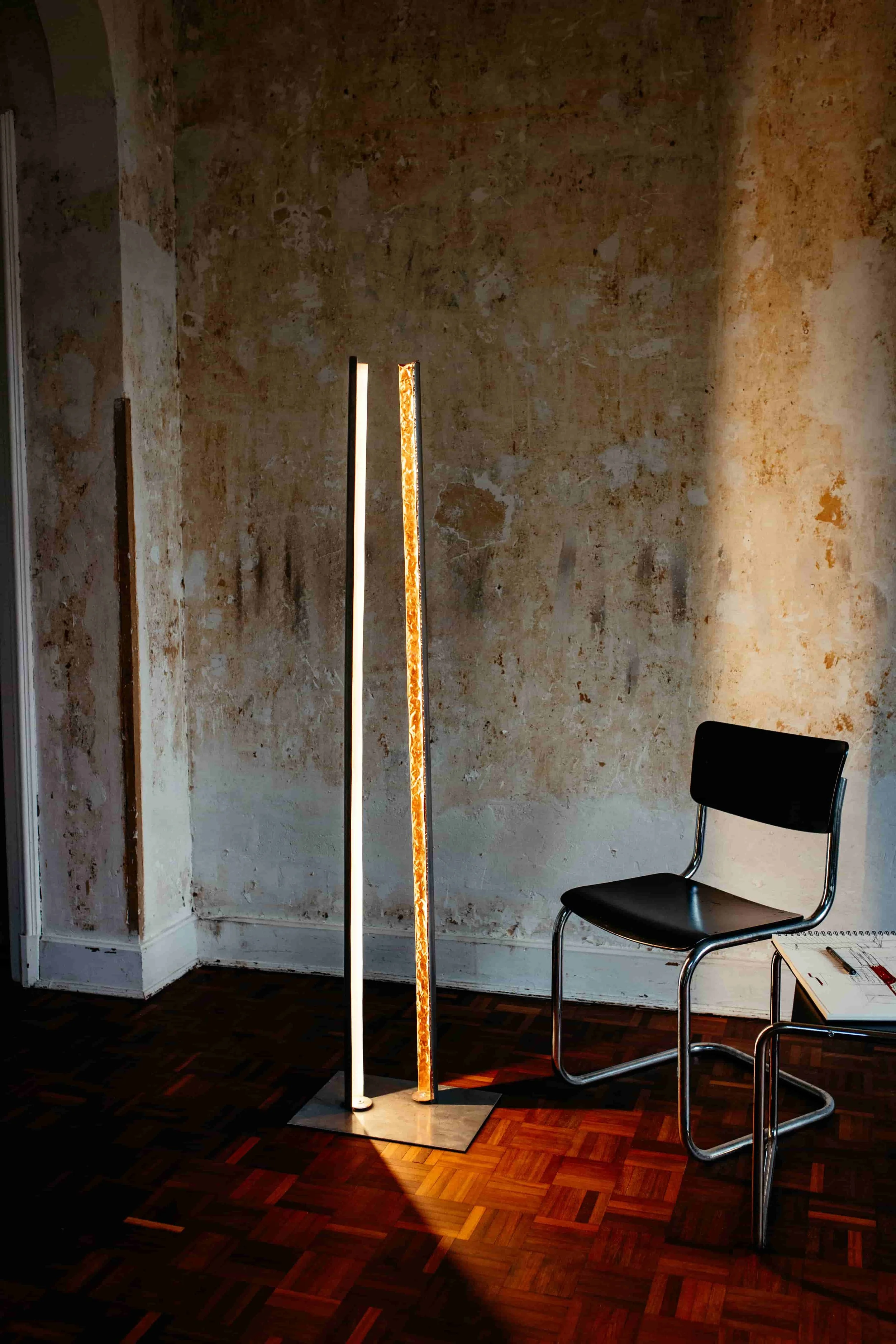 Zwei beleuchtete Lichtleisten vor einer alten, verputzten Wand, daneben ein schwarzer Stuhl.