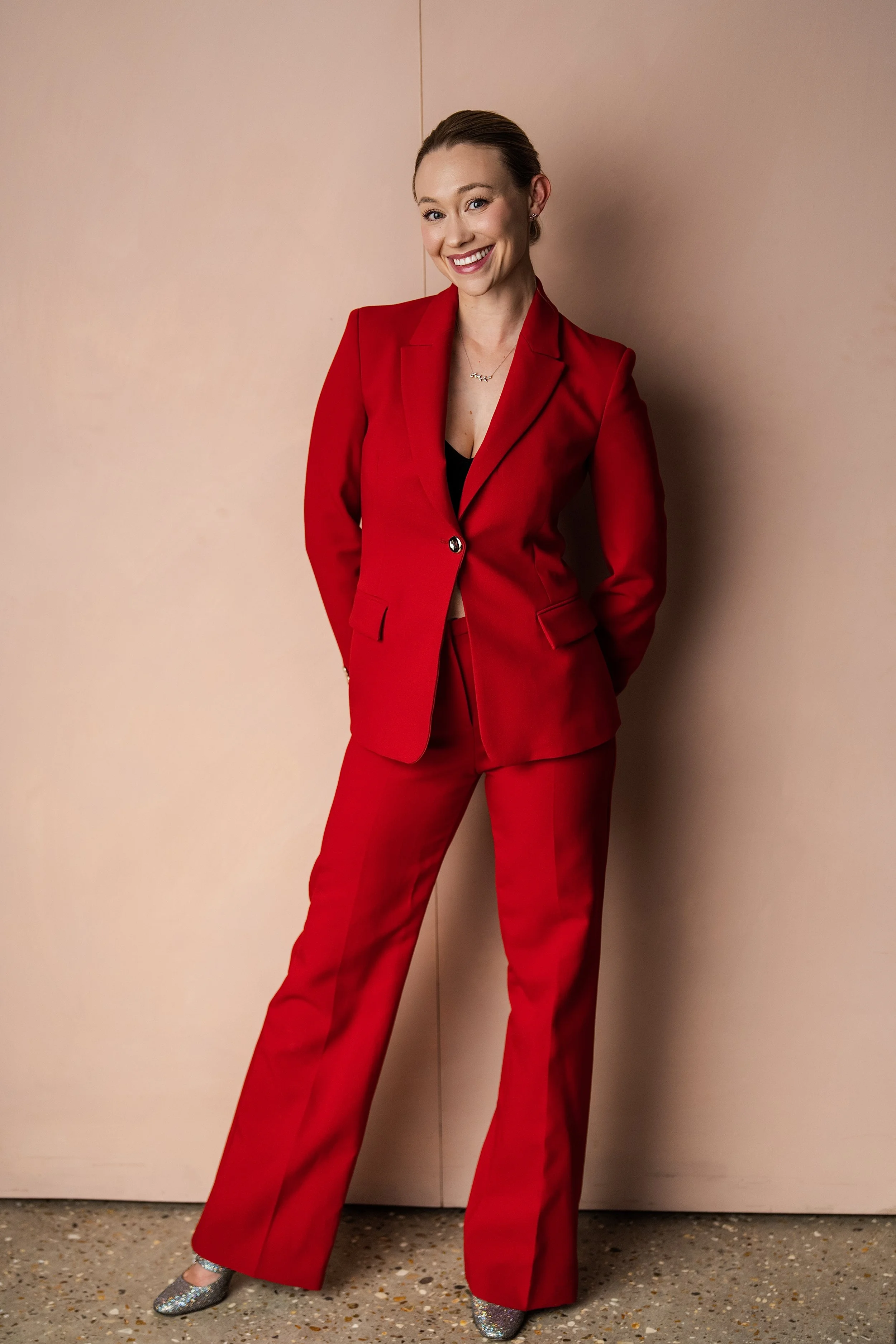 Amy Whitworth dressed in a red suit with matching pants, standing against a plain wall, smiling at the camera, wearing sparkly silver shoes, and a necklace. Photo by Yellowbelly.