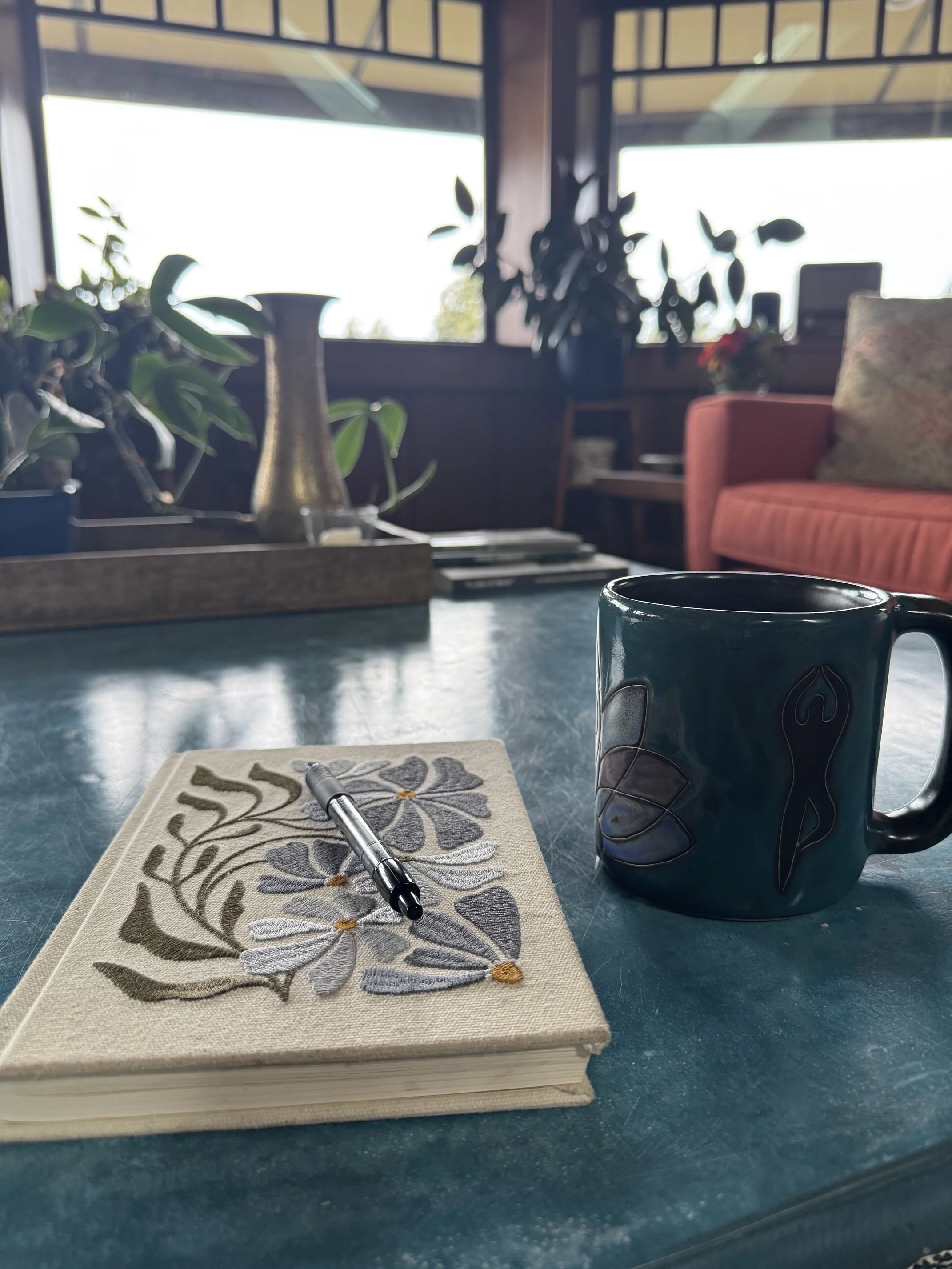 Close-up of a closed beige notebook with a floral pattern on a table, a black pen rests on top, and a dark blue mug with a flame design is in the foreground. In the background, there are houseplants, a bookshelf, and a cozy living room with large windows letting in natural light.