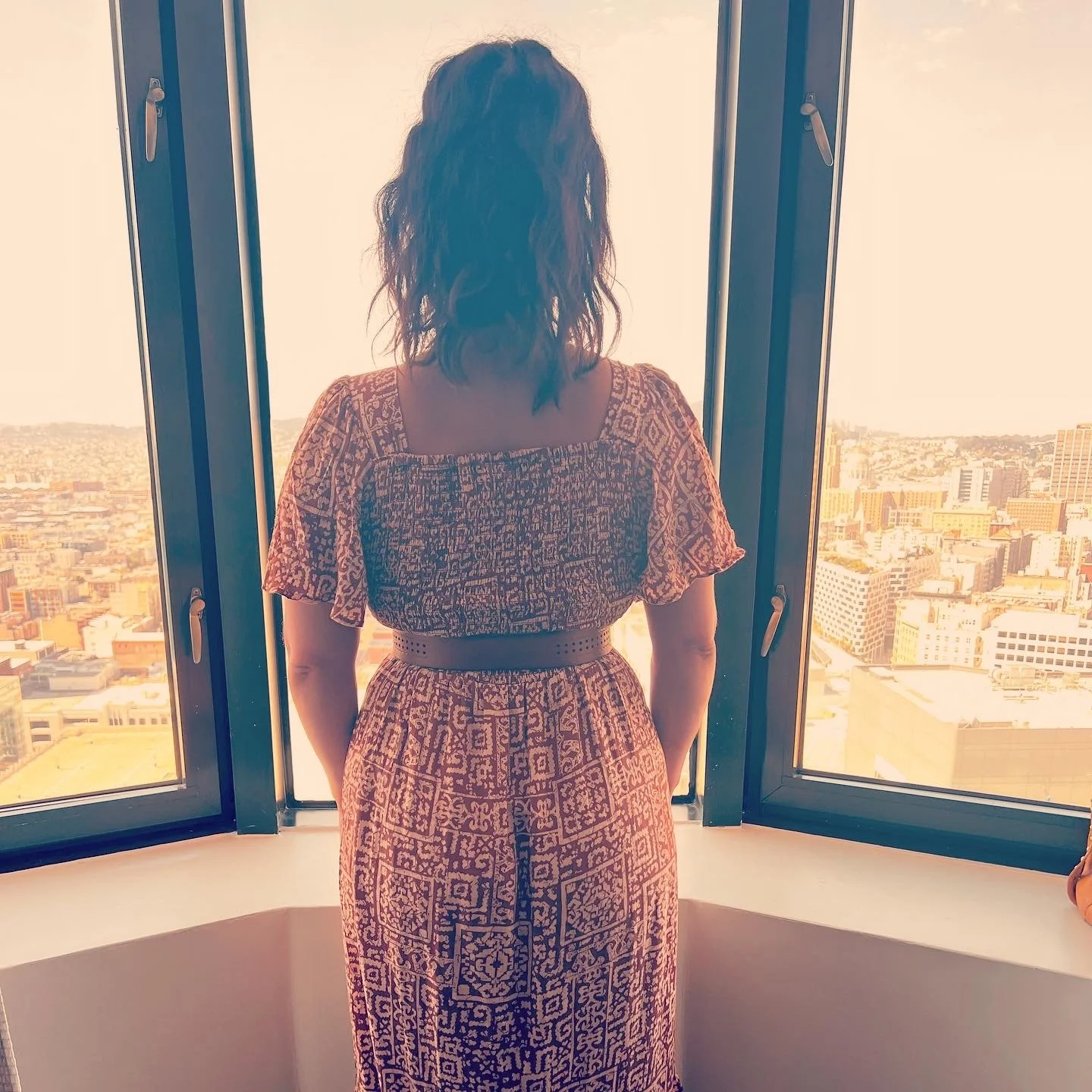 A woman with dark, wavy hair standing indoors, looking out of large windows at a cityscape with tall buildings under a clear sky.