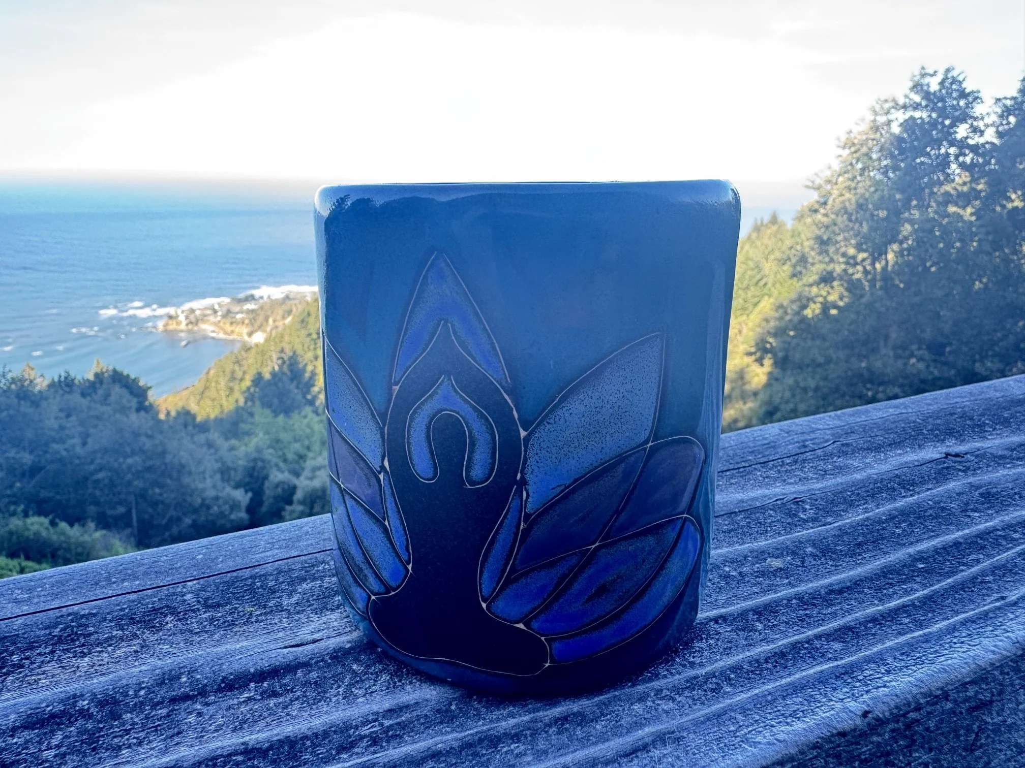 A blue ceramic mug with a carved design, placed on a wooden surface overlooking a scenic ocean view with green hills and a distant coastline.