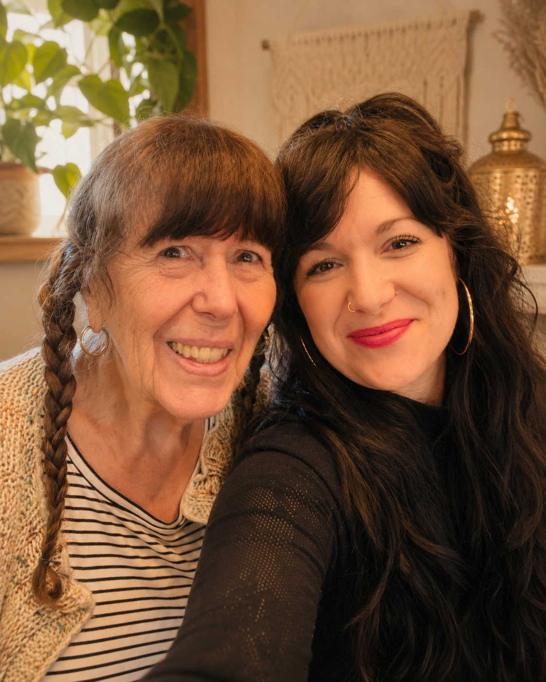 Two women smiling closely for a selfie indoors, with potted plants and decorative items in the background.