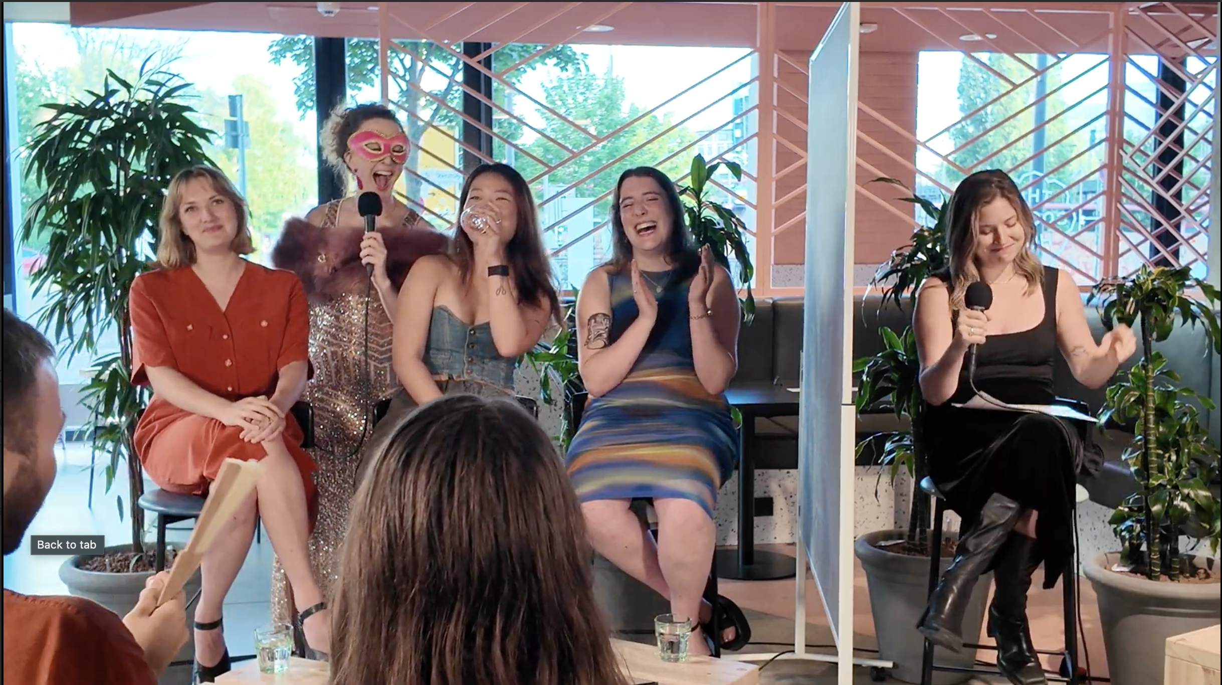 Five women on stage participating in a panel or discussion, with one woman wearing a pink mask and holding a microphone, laughing and clapping, in a room with large windows and plants.