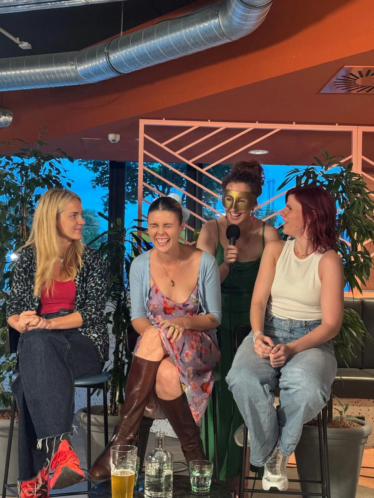 Four women sitting and one woman standing, holding a microphone, in a social setting with plants and modern decor.