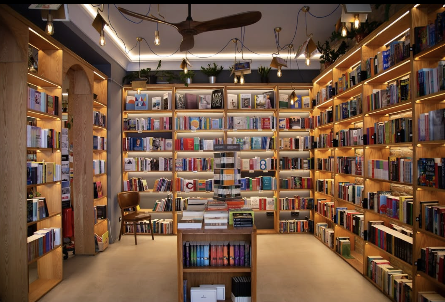 Interior of a bookstore with wooden shelves filled with books, a small table with books, a chair, plants on top of shelves, and decorative hanging lights.