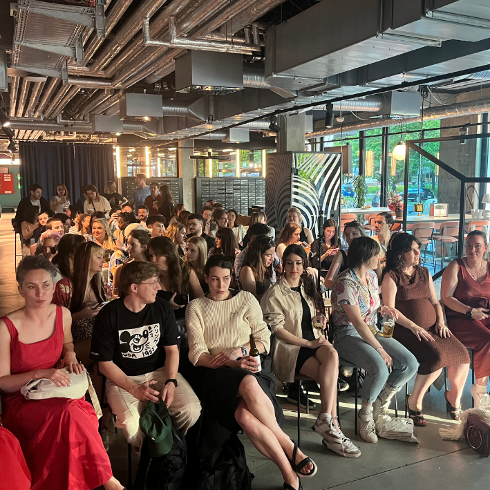 A large group of people seated indoors during an event, with some holding drinks, in a modern space with industrial ceiling and large windows.