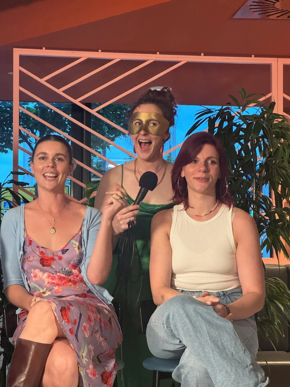 Three women sitting together indoors, one holding a microphone, with plants and a decorative background behind them.