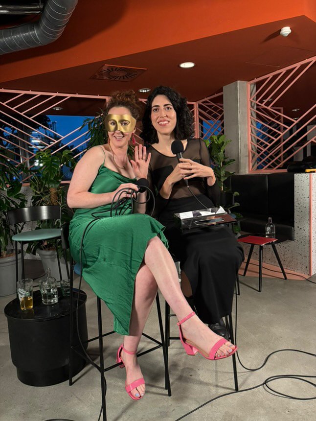 Two women sitting on high chairs at a social event, holding microphones. One woman is wearing a green dress, pink heels, and a gold mask, waving and smiling. The other woman is in a black dress, holding a microphone, smiling. There are drinks on a sm