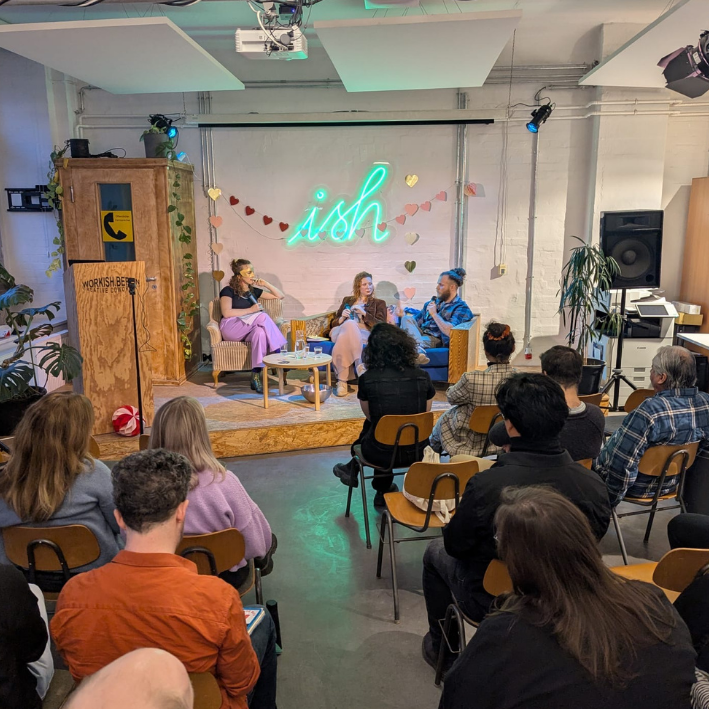 Panel discussion at a small event with three speakers on a stage decorated with a neon sign saying 'ish' and heart-shaped decorations, with an audience seated in chairs watching.