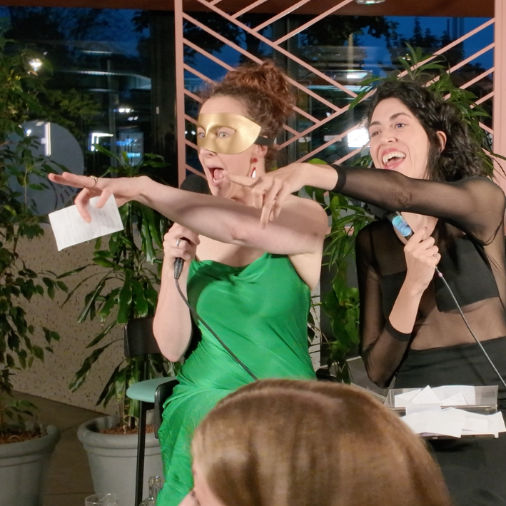 Two women performing comedy or entertainment act, one wearing a green dress and a gold mask, holding a microphone and pointing, the other in a black sheer top, also holding a microphone and laughing. They are on a stage with plants and a decorative b