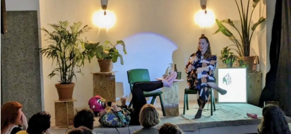 A woman sitting on a stage with two potted plants, addressing an audience. She is wearing a patterned dress and holding a microphone. There is a person lying on the floor in front of her, wearing colorful clothing and pink shoes, taking a photo or re