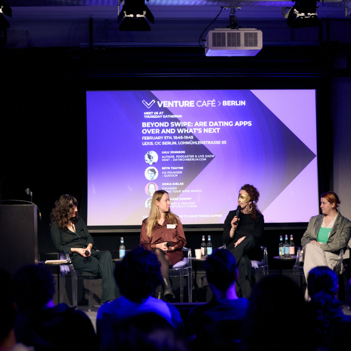 A panel discussion at an event called Venture Café Berlin with four women speakers and an audience. The background screen displays event details about dating apps and entrepreneurship.