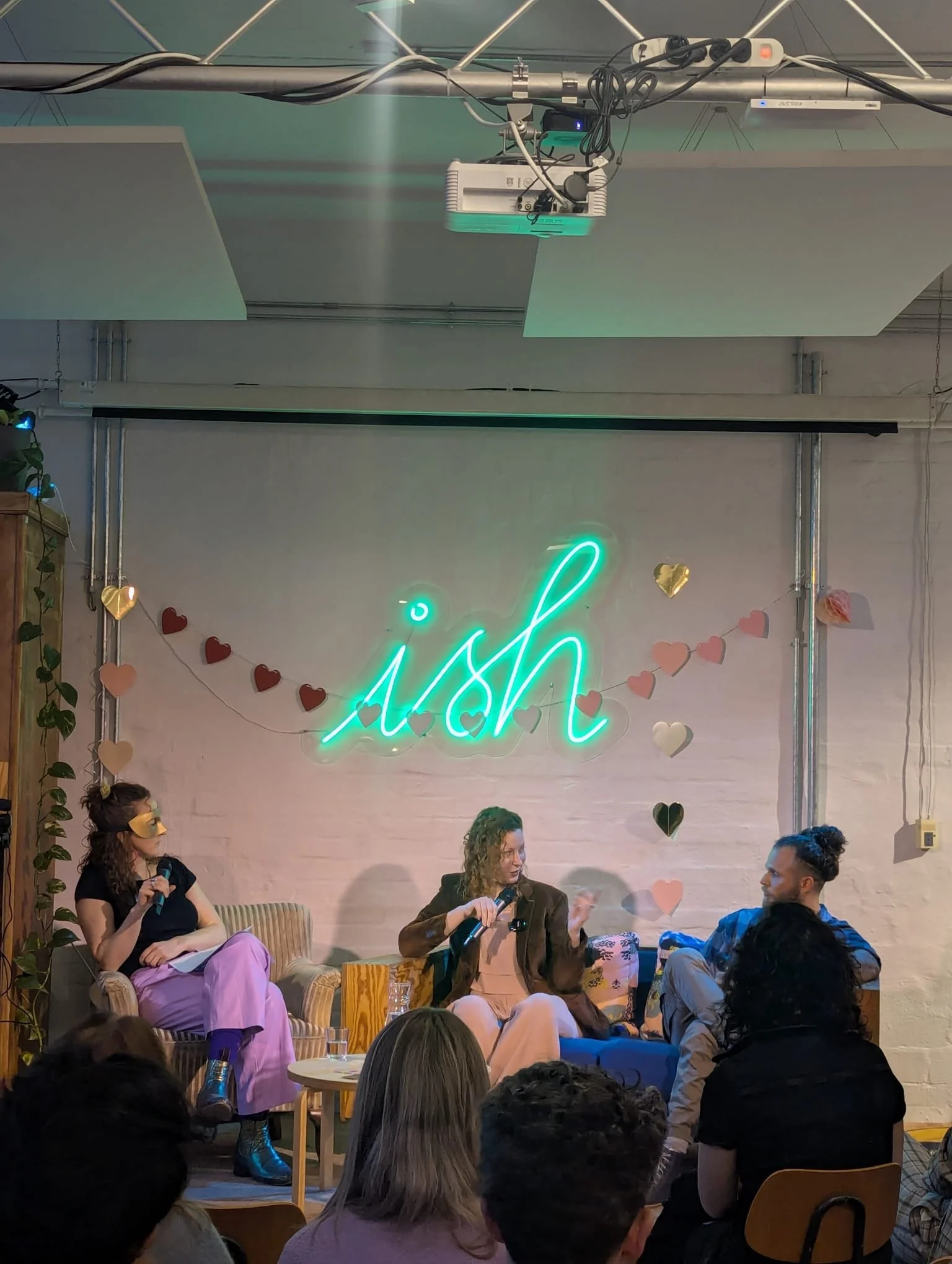 A panel discussion with three women sitting on a stage with a neon sign that says 'ish' behind them, decorated with paper hearts and fairy lights, in a cozy, casual setting.