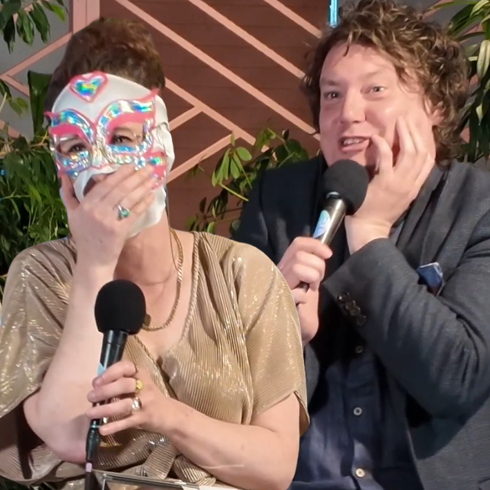 Two people sitting at a table, one wearing a decorative mask and the other speaking into a microphone, with plants and a wooden lattice background.