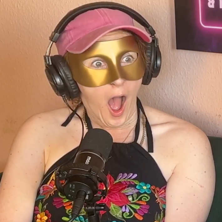 A woman wearing headphones, a pink hat, and a gold mask, with her mouth open in surprise, speaking into a microphone.