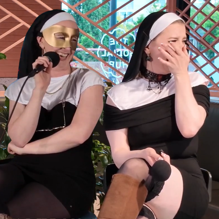 Two women dressed as nuns, both wearing black and white habits, sitting next to each other; one with a gold mask and holding a microphone, smiling, and the other covering her face, also holding a microphone, in a modern indoor setting with wooden dec