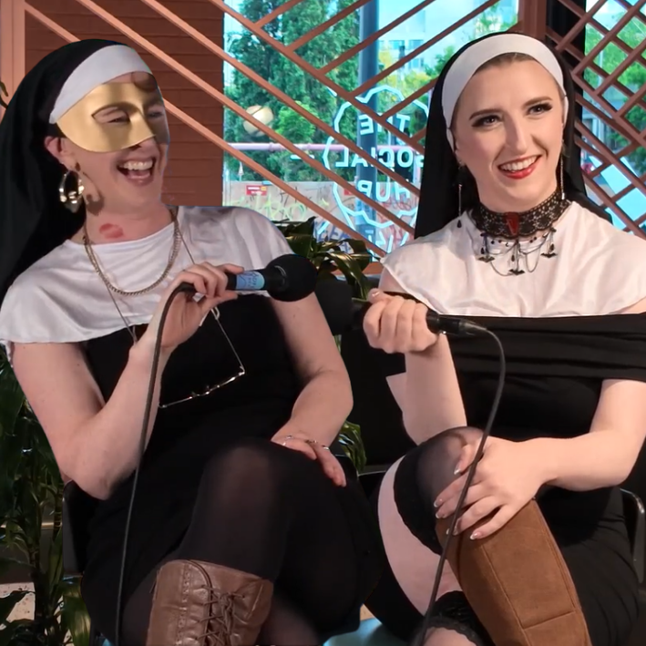 Two women dressed as nuns with humorous masks, sitting and laughing while holding microphones in a setting with a decorated backdrop and plants.