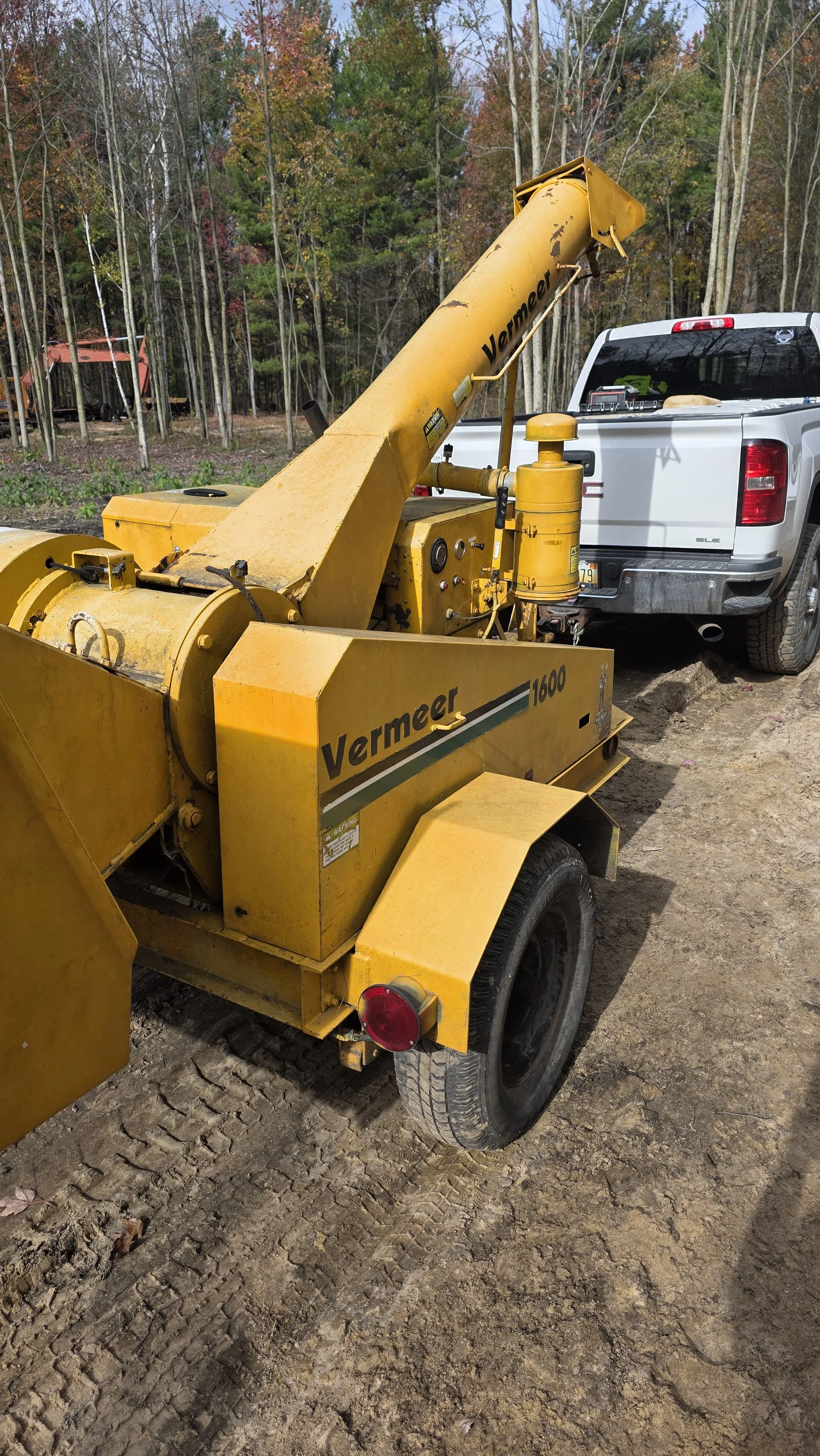 Yellow Vermeer trencher attached to a white pickup truck, parked on a dirt surface with trees in the background.